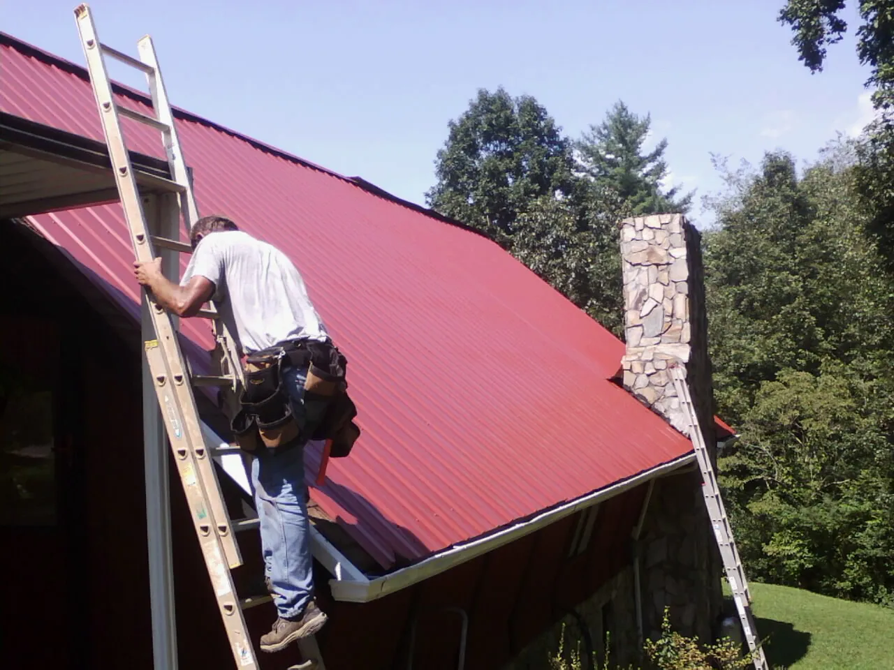 Roofer on a ladder beside a red metal roof and stone chimney — Soffit & Fascia Repair by Roofing Brothers in Fisher